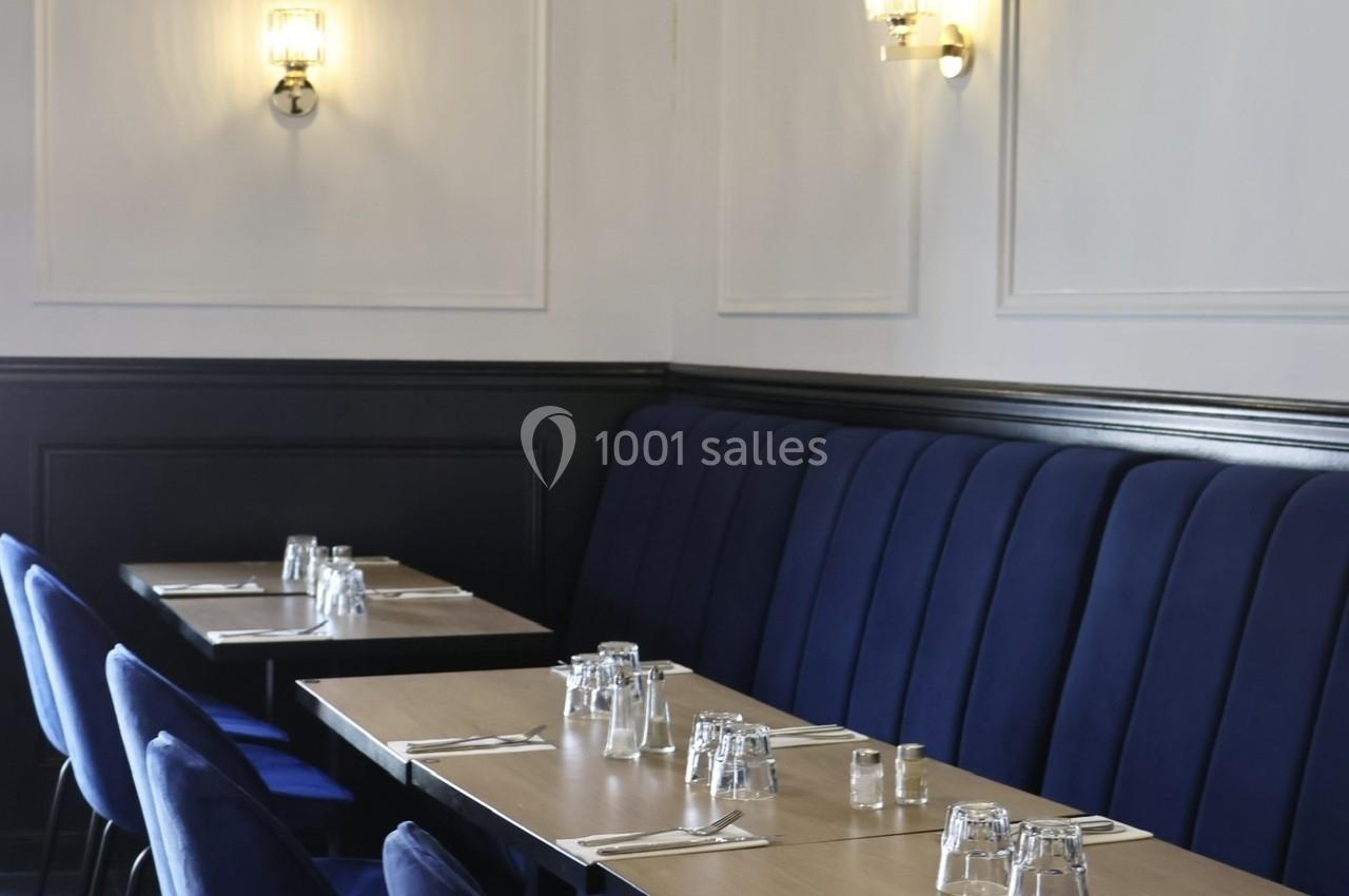 Salle de restaurant avec banquettes bleues, tables en bois clair dressées avec verres et serviettes, éclairage mural.