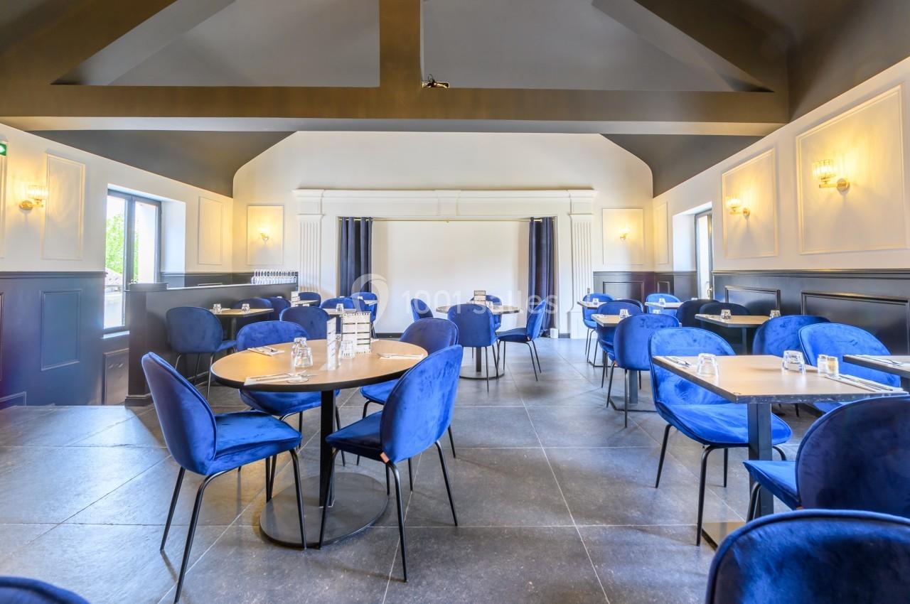 Salle de restaurant lumineuse avec des tables rondes, des chaises bleues et un plafond aux poutres apparentes.