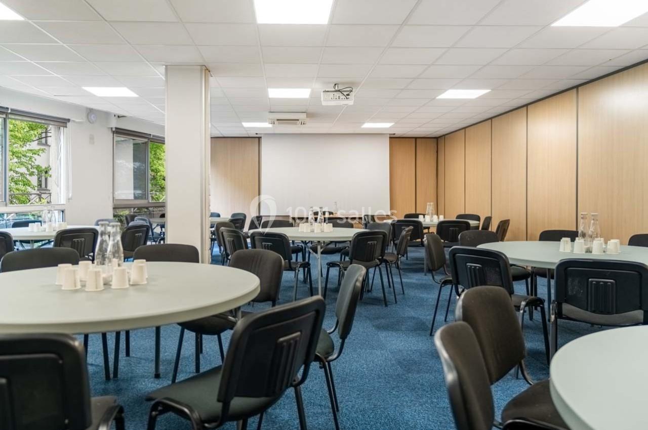 Salle de réunion avec tables rondes, chaises noires, lumière naturelle et projecteur au plafond.