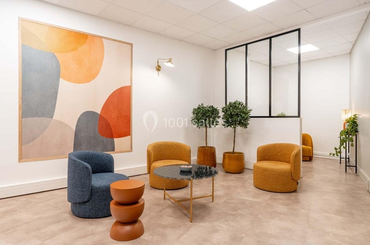 Espace lumineux avec fauteuils colorés, table basse en verre, plantes en pot et une grande œuvre murale abstraite.