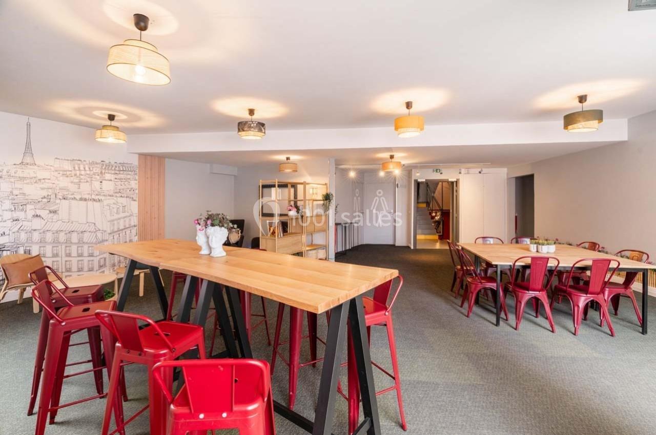 Salle lumineuse avec tables en bois, chaises rouges, décoration murale de Paris et éclairage suspendu.