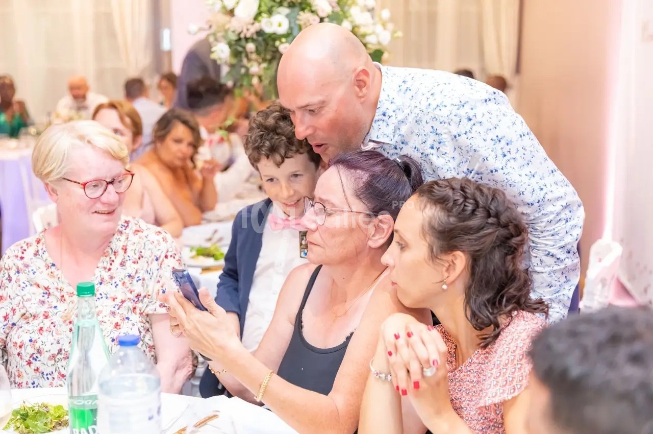 Un groupe de personnes assises autour d'une table, regardant un téléphone, dans un cadre festif avec des fleurs en arrière…