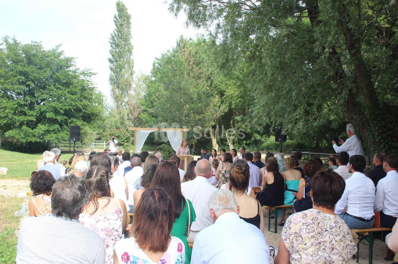 Un groupe de personnes assiste à une cérémonie en plein air, sous des arbres, avec un officiant devant une arche décorée.