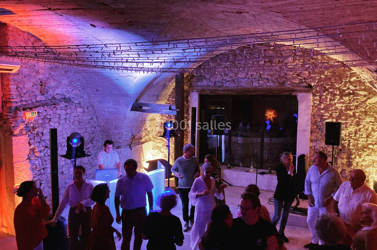 Personnes dansant et discutant dans une salle voûtée en pierre, éclairée par des lumières colorées, avec un DJ en arrière…