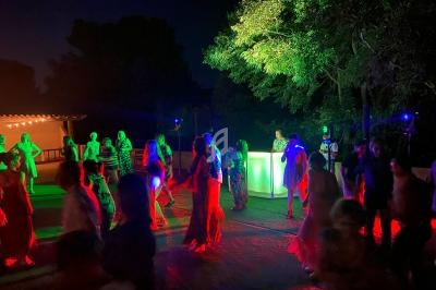 Des personnes dansent en extérieur la nuit, éclairées par des lumières colorées et entourées d'arbres.