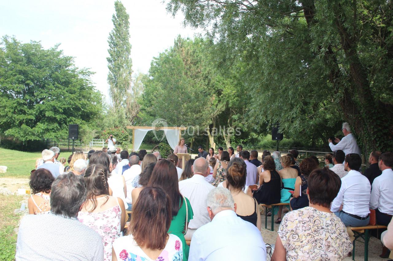 Un groupe de personnes assiste à une cérémonie en plein air sous des arbres, avec une arche décorée en arrière-plan.