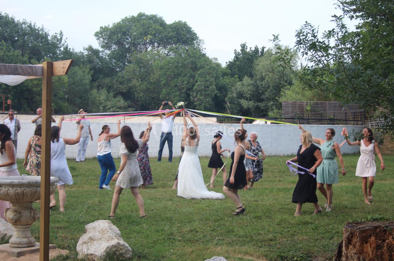 Des personnes dansent en cercle autour d'une mariée tenant des rubans colorés, dans un jardin en plein air.