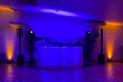 Console de DJ éclairée en violet et jaune, entourée de plantes et d'équipements audio dans une salle sombre.