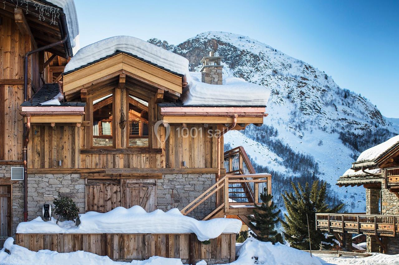 Chalet en bois entouré de neige avec une montagne en arrière-plan sous un ciel bleu clair.