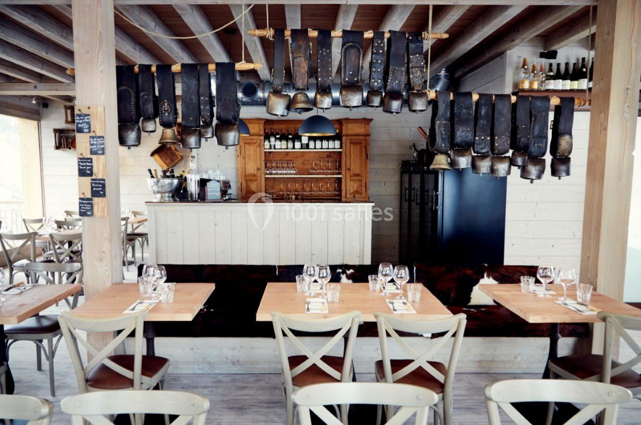 Salle de restaurant rustique avec tables en bois, chaises croisées et décoration de cloches suspendues au plafond.