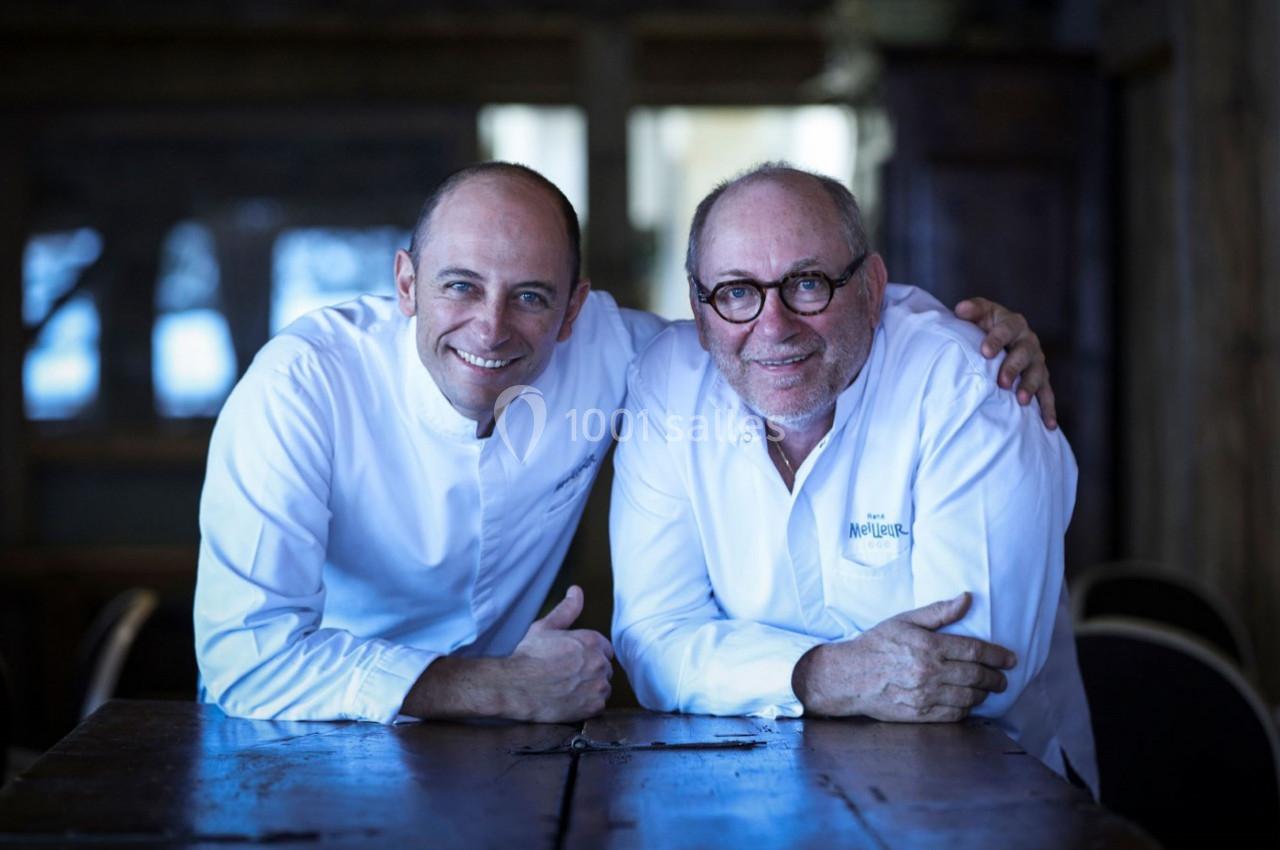 Deux chefs en tenue blanche sourient et posent côte à côte, assis à une table en bois dans un intérieur sombre.