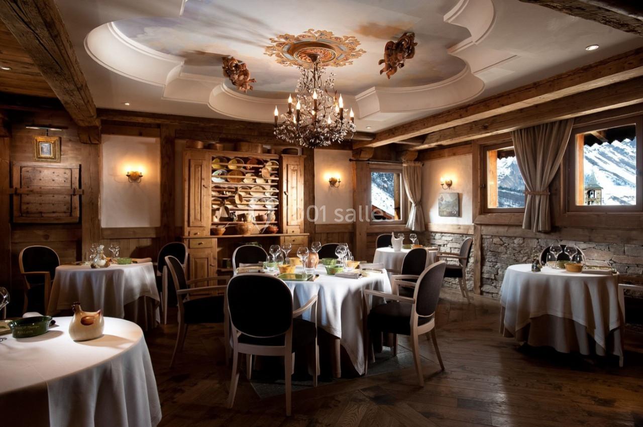 Salle de restaurant élégante avec tables dressées, décor en bois, lustre central et vue sur des montagnes enneigées.