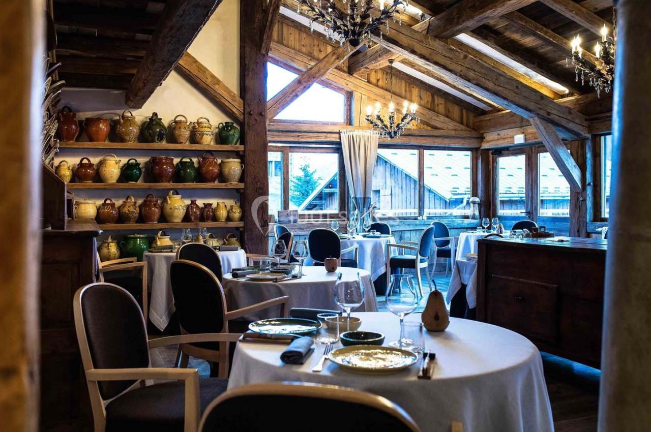 Salle de restaurant chaleureuse avec poutres apparentes, tables dressées et étagères décorées de pots en céramique.