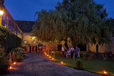 Entrée en pierre éclairée de nuit, entourée de fleurs et d'arbres, avec un portail en fer forgé.
