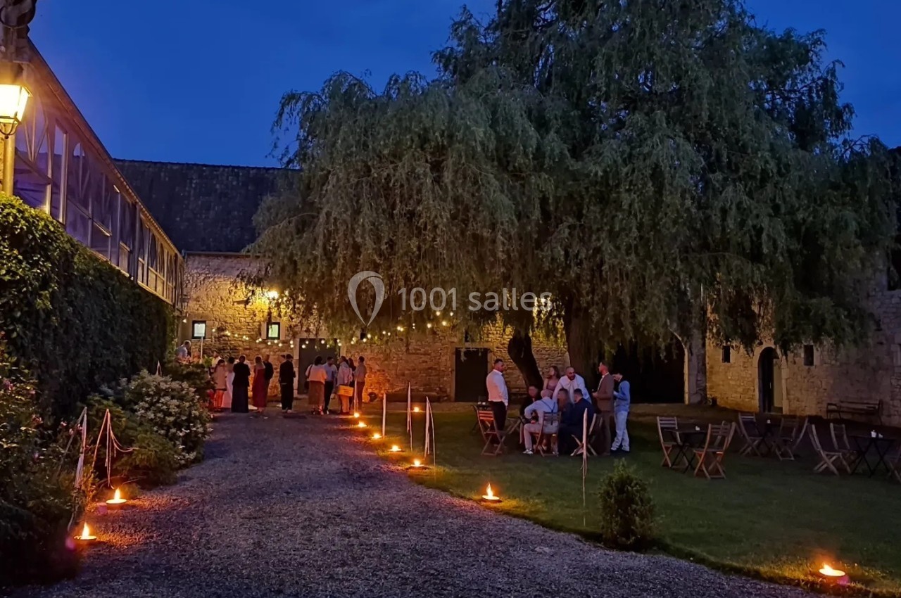 Cour éclairée par des lanternes au crépuscule, avec des invités rassemblés près d'un bâtiment en pierre et un grand arbre.