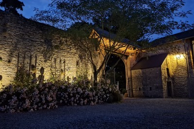 Entrée en pierre éclairée de nuit, entourée de fleurs et d'arbres, avec un portail en fer forgé.
