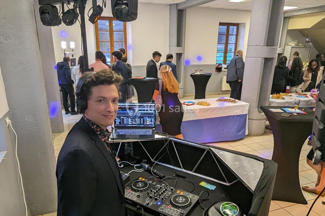 Un DJ anime un événement dans une salle avec des invités debout près de tables hautes et des buffets.