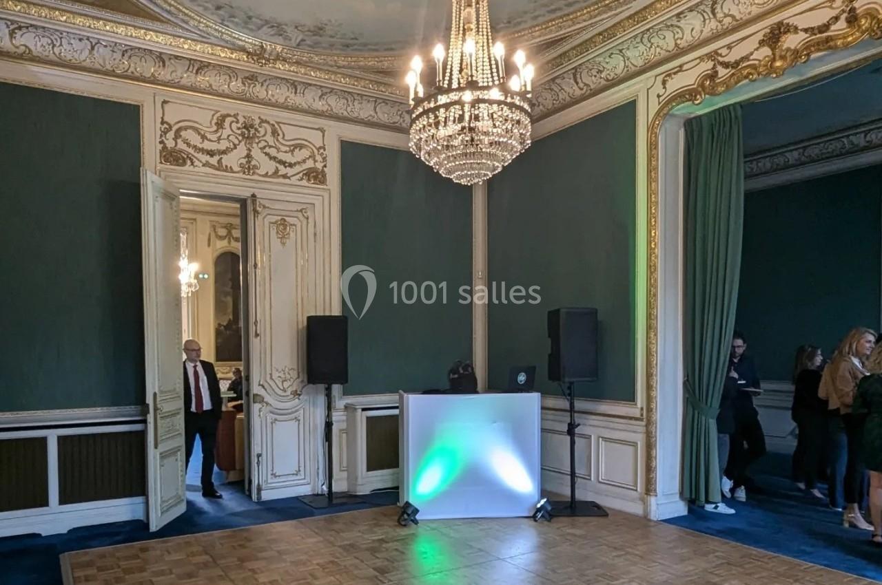 Salle élégante avec moulures dorées, murs verts, parquet, lustre central et équipement audio installé près d'une table…