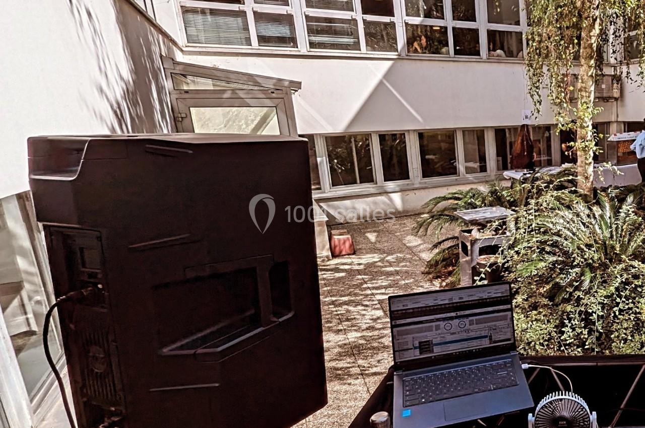 Poste de DJ en extérieur avec ordinateur portable, enceinte et végétation, devant un bâtiment moderne avec grandes fenêtres.