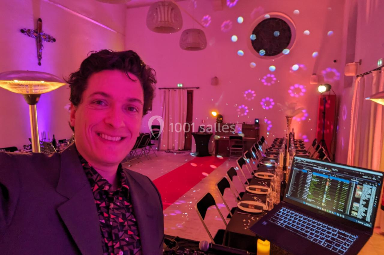 Un homme souriant dans une salle décorée avec des lumières colorées, devant une table de mixage et un ordinateur portable.