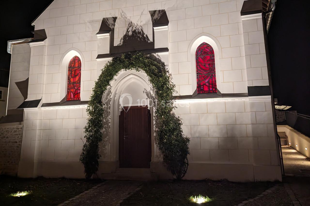 Façade d'une petite chapelle éclairée de nuit, avec une porte en bois entourée de verdure et des vitraux rouges.
