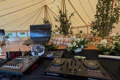 Table de mixage et ordinateur installés dans une tente décorée pour un événement avec des fleurs et des tables dressées.