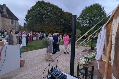 Un groupe de personnes rassemblées en extérieur près d'une tente éclairée, dans un cadre champêtre avec arbres et pelouse.