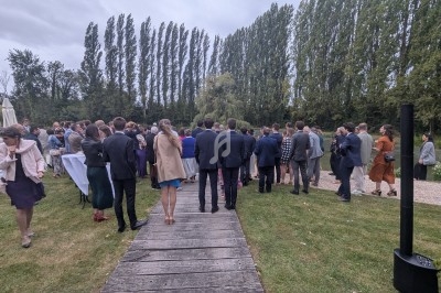 Groupe de personnes rassemblées en extérieur sur une pelouse près d'un chemin en bois, entouré d'arbres.
