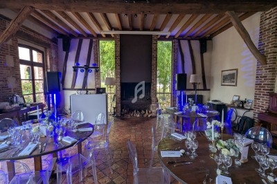 Salle de réception avec tables dressées, chaises transparentes, éclairage bleu et cheminée en fond.