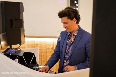 Un DJ en costume bleu mixe de la musique sur une table de mixage dans un espace intérieur éclairé.
