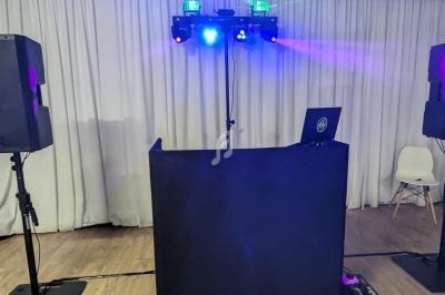 Poste de DJ avec table noire, ordinateur portable, enceintes et jeux de lumières colorées dans une salle avec rideaux blancs.