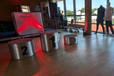 Podium métallique numéroté 1, 2, 3 dans une salle avec parquet, éclairage rouge et vue sur une piste extérieure.