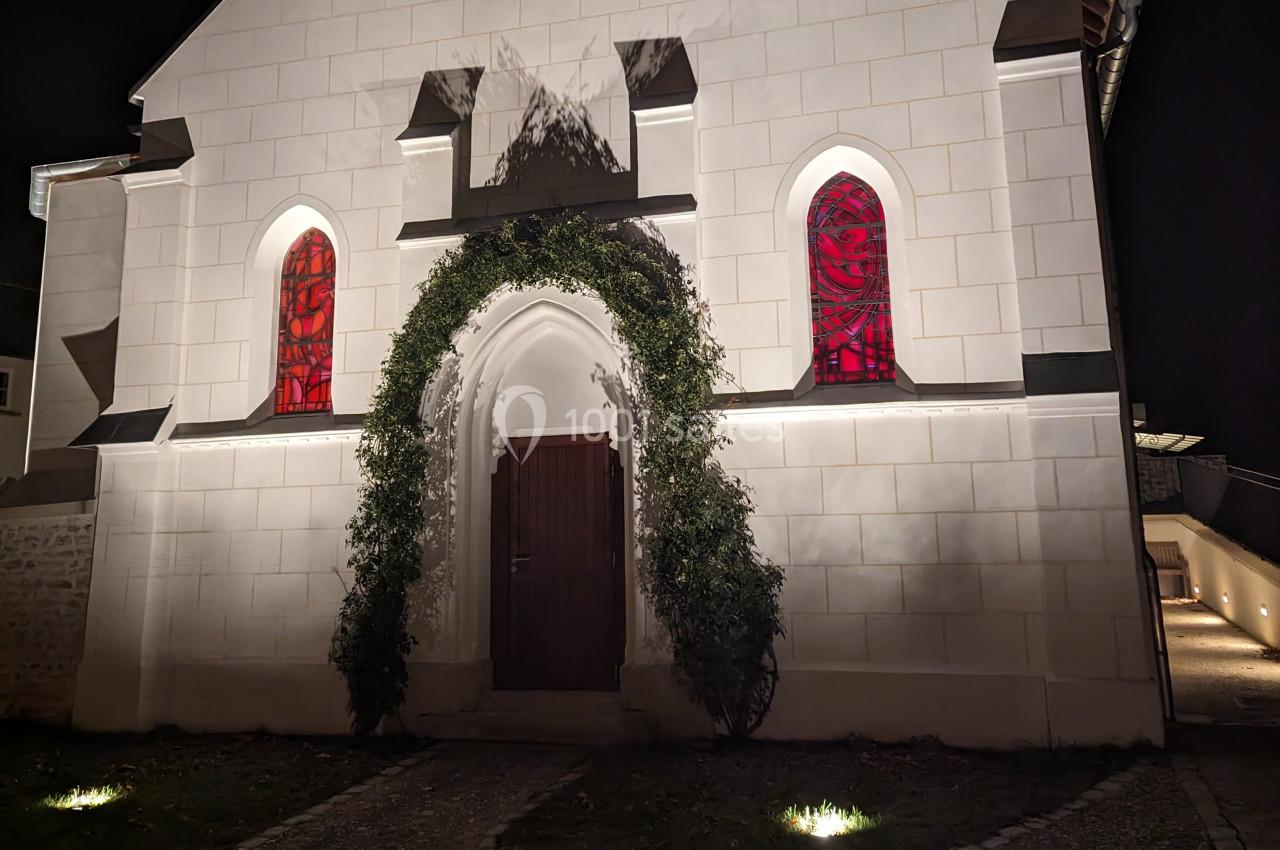 Façade d'une petite chapelle blanche éclairée de nuit, avec une porte en bois entourée de verdure et des vitraux rouges.