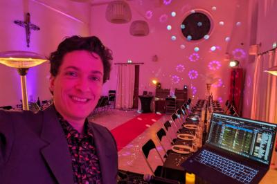 Un homme s'exprime au micro devant un public rassemblé dans une cour extérieure entourée de végétation.