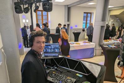 Un homme s'exprime au micro devant un public rassemblé dans une cour extérieure entourée de végétation.