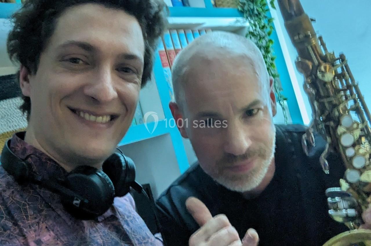 Deux hommes posent ensemble, l'un tenant un saxophone, dans un environnement intérieur avec des étagères en arrière-plan.