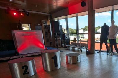 Un homme s'exprime au micro devant un public rassemblé dans une cour extérieure entourée de végétation.