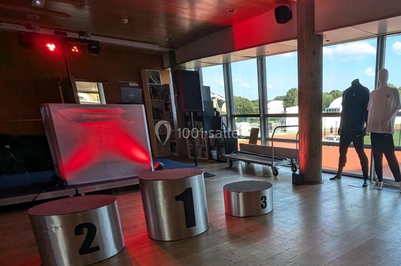 Podium métallique numéroté dans une salle lumineuse avec baies vitrées donnant sur une piste extérieure.