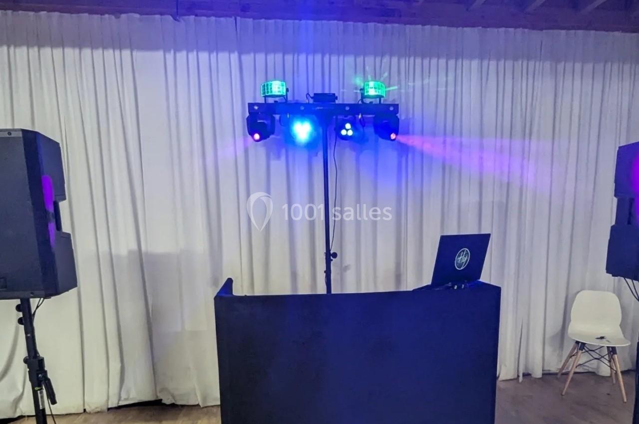 Poste de DJ avec table noire, ordinateur portable, enceintes et jeux de lumières colorées devant un rideau blanc.