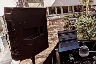 Un homme s'exprime au micro devant un public rassemblé dans une cour extérieure entourée de végétation.