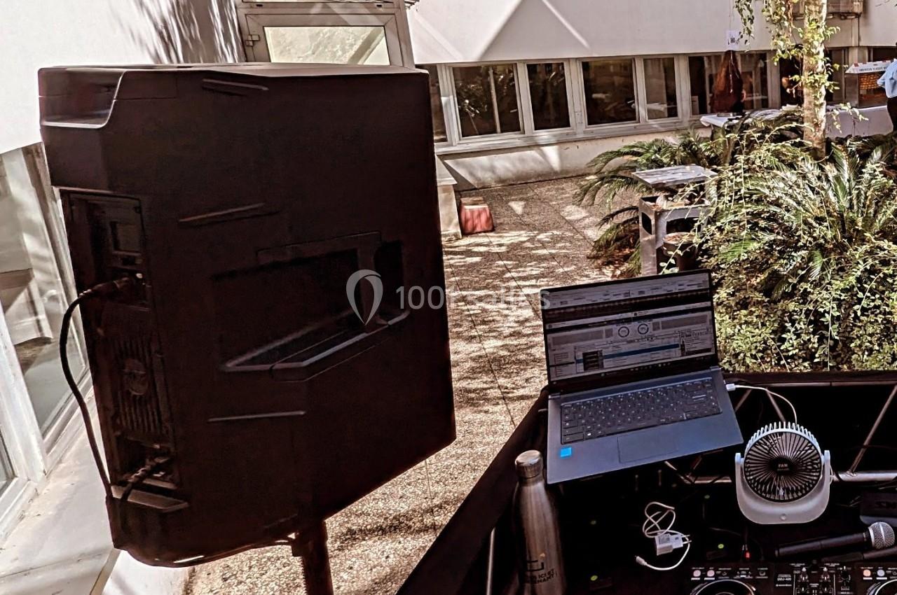 Poste de DJ en extérieur avec ordinateur portable, enceinte, ventilateur et plantes en arrière-plan.