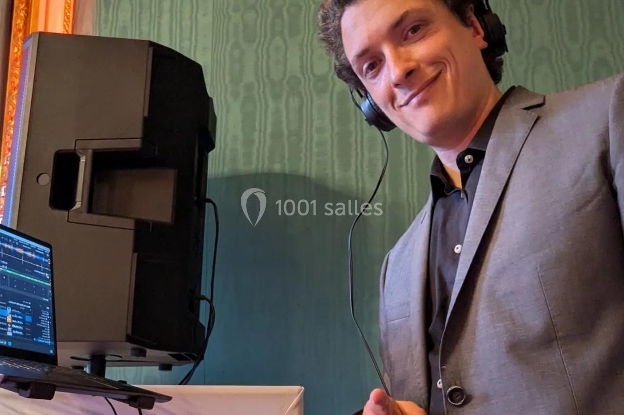 Un DJ souriant en costume mixe de la musique avec un casque, devant une enceinte et un ordinateur portable.