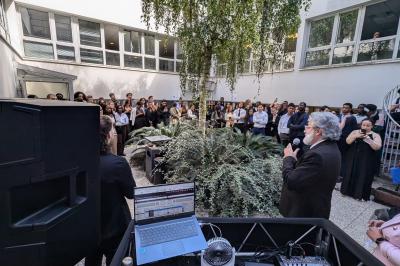 Un homme s'exprime au micro devant un public rassemblé dans une cour extérieure entourée de végétation.