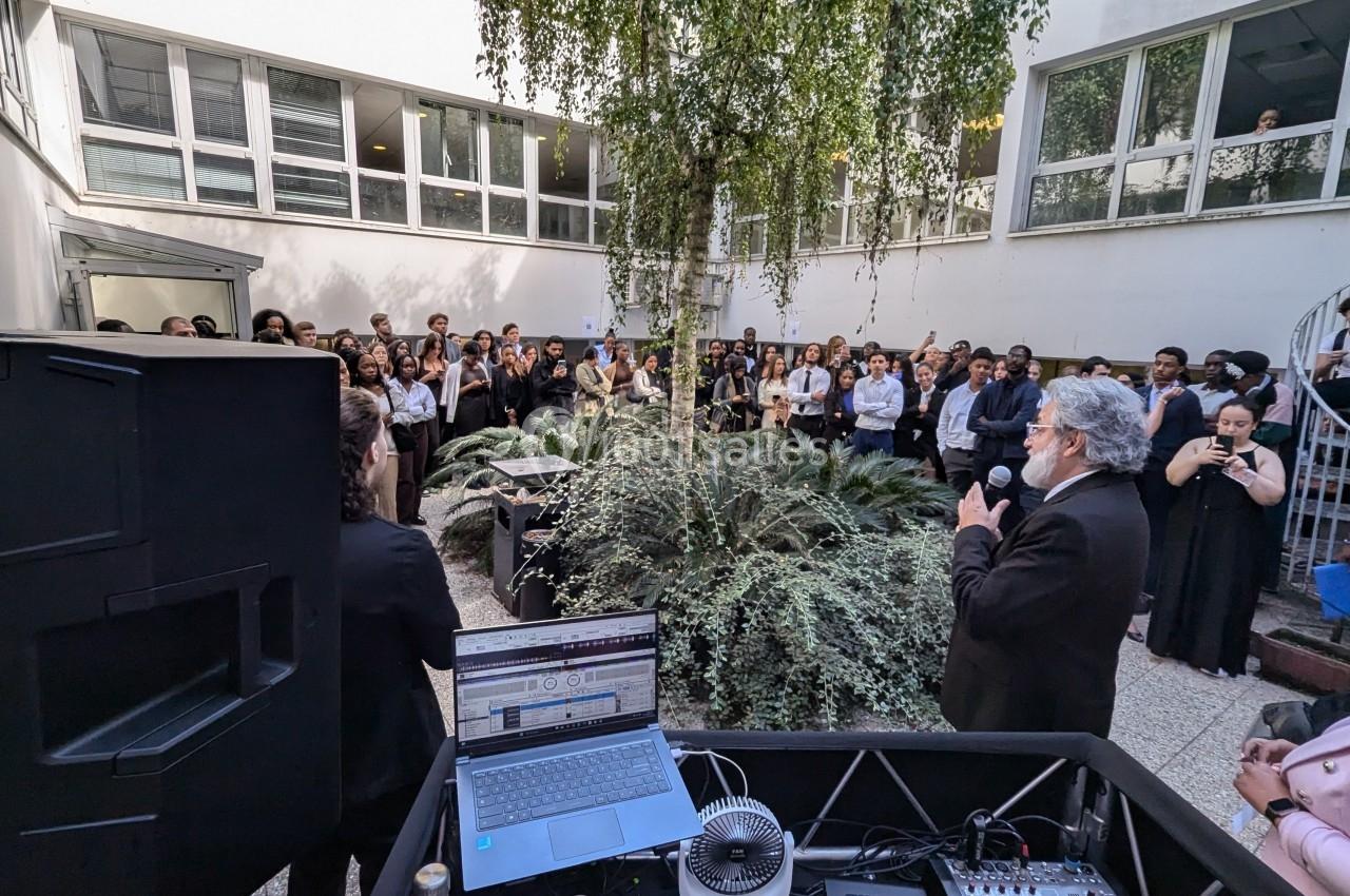 Un homme s'exprime au micro devant un public rassemblé dans une cour extérieure entourée de végétation.