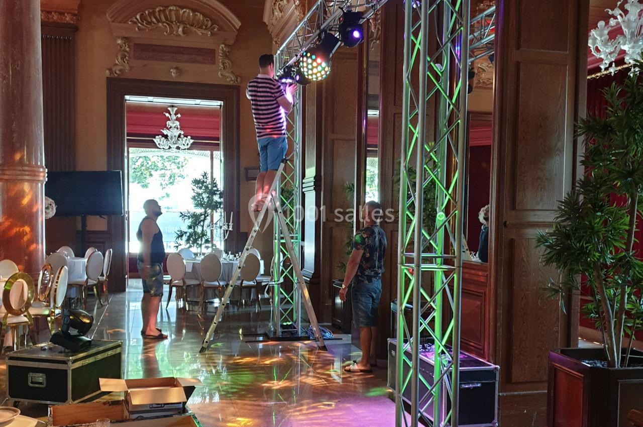 Un technicien ajuste un projecteur sur une structure métallique dans une salle élégante avec des tables et des lustres.