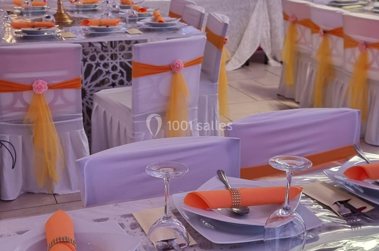 Tables décorées pour un événement avec nappes blanches, serviettes orange, chaises ornées de rubans jaunes et roses.