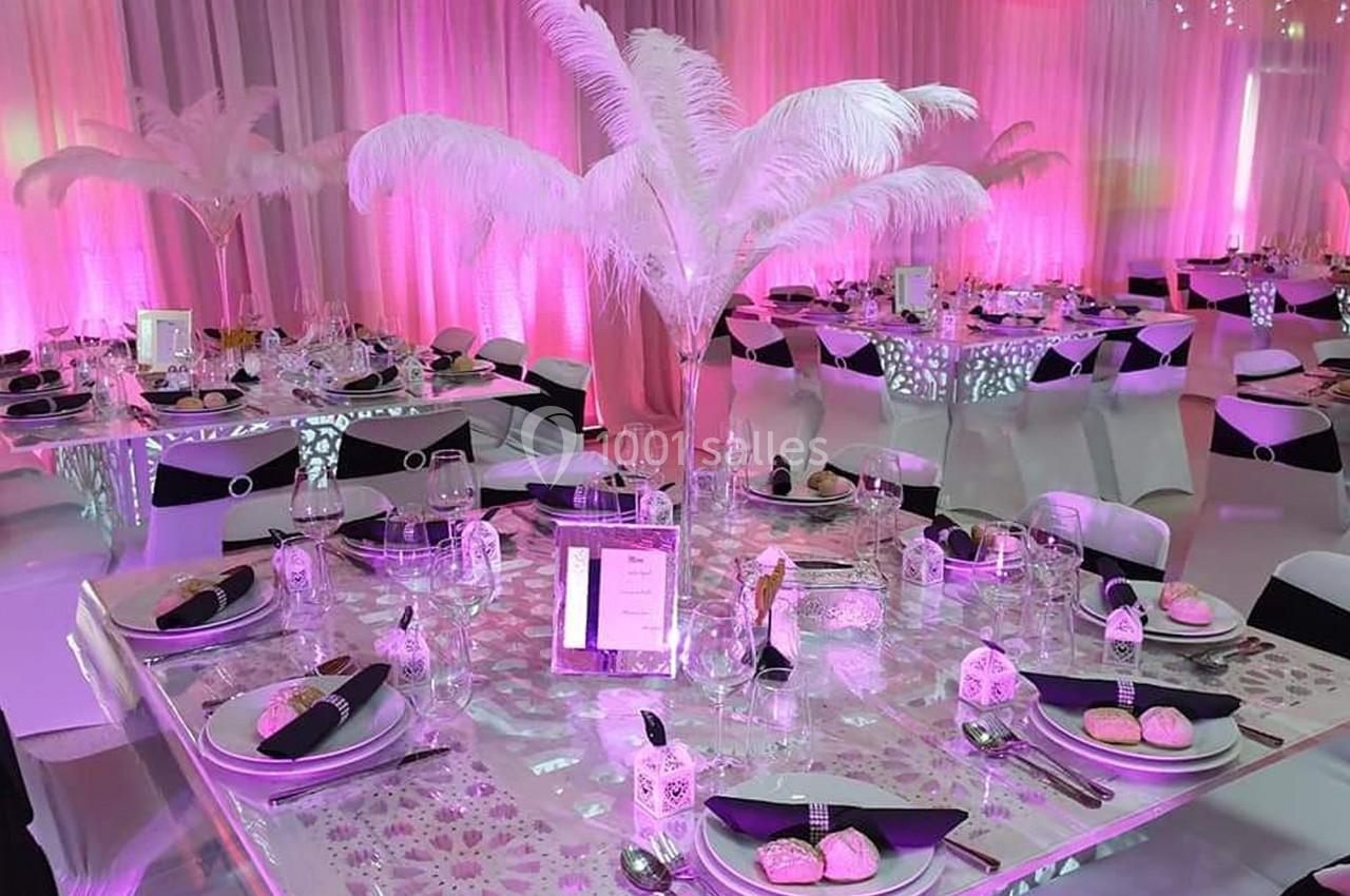 Salle de réception décorée avec des tables élégantes, nappes scintillantes, plumes blanches et éclairage rose.