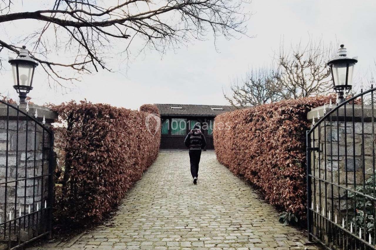 Un homme marche sur une allée pavée bordée de haies rouges menant à une maison en arrière-plan.