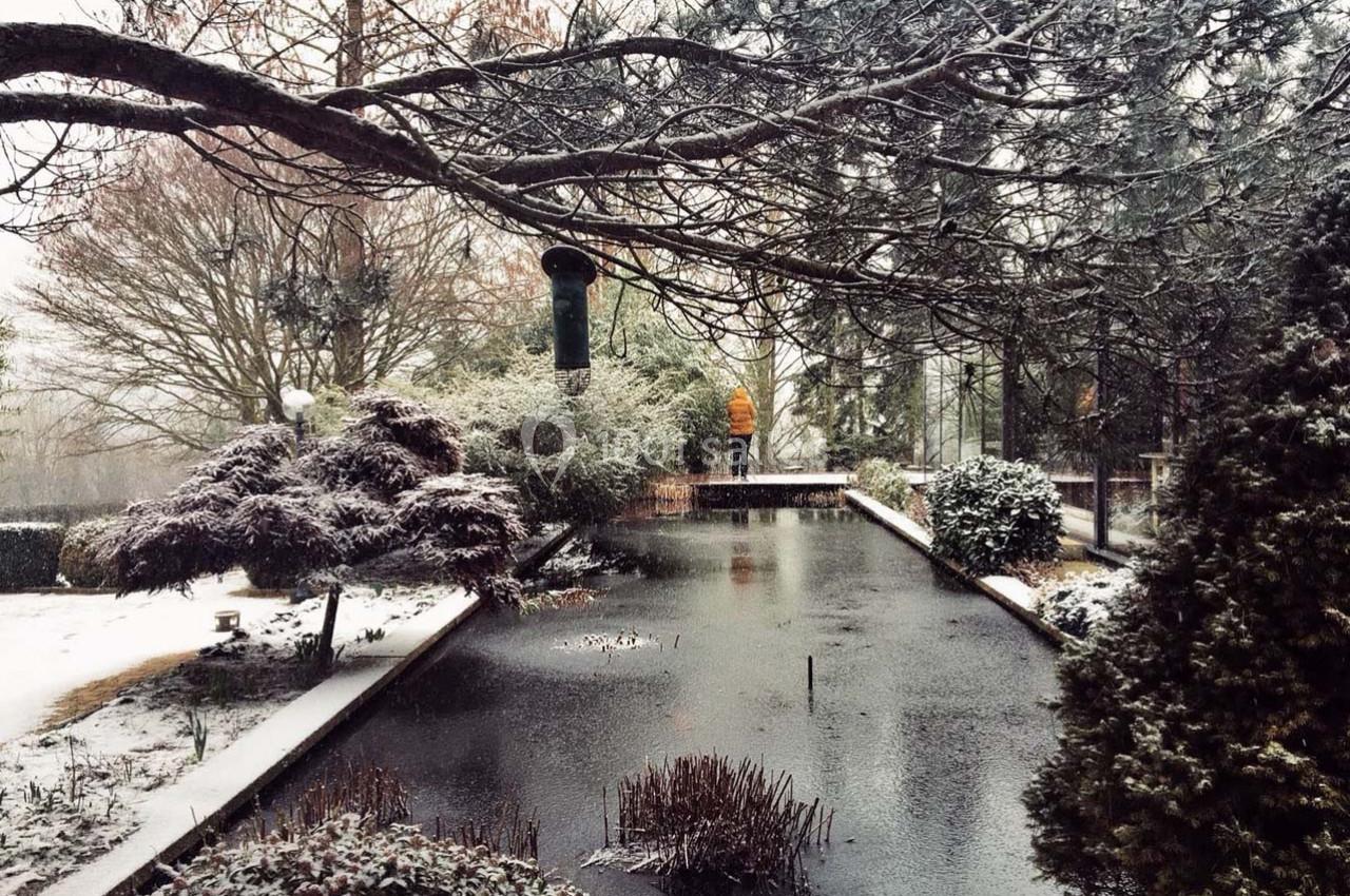 Un jardin enneigé avec un étang gelé, des arbres dénudés et une personne en manteau jaune au loin.