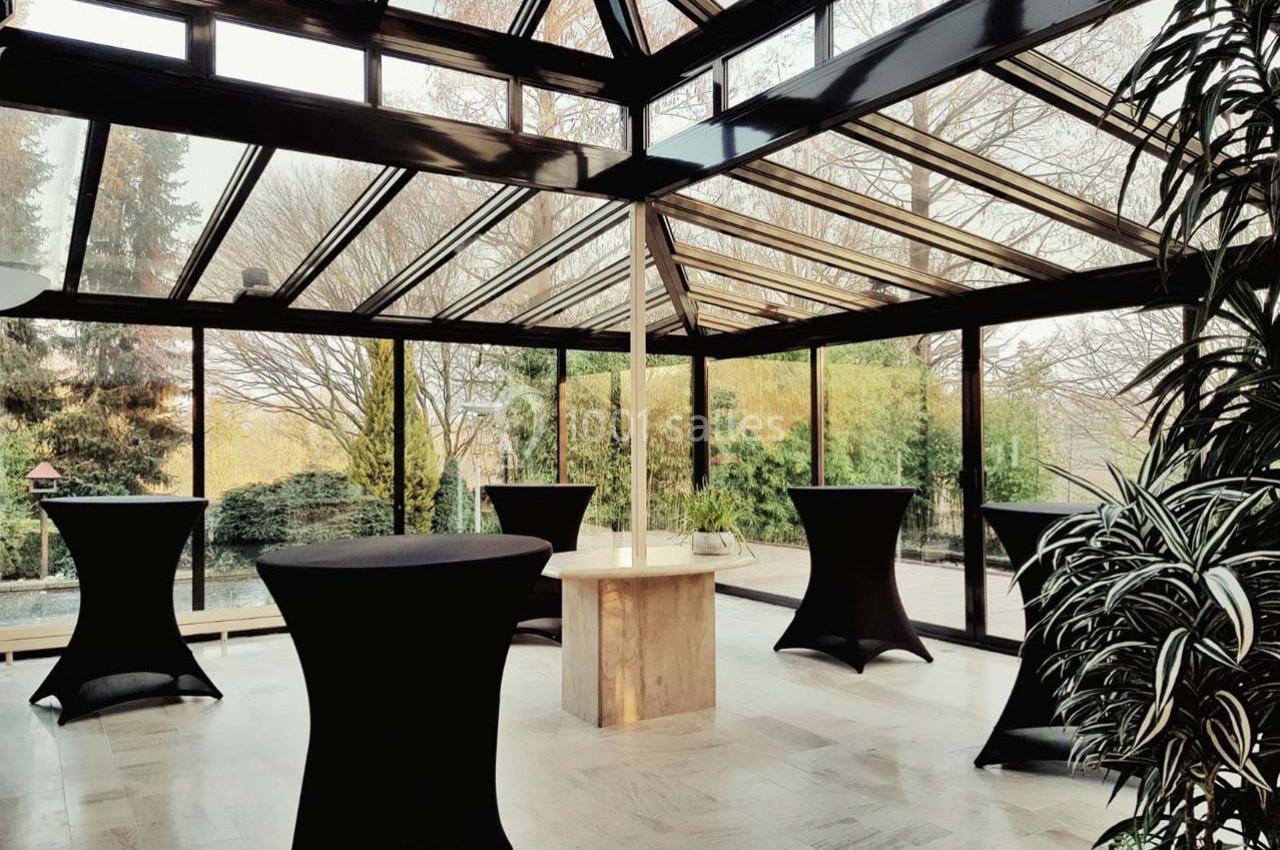 Salle lumineuse avec verrière, tables hautes drapées de noir et vue sur un jardin arboré en arrière-plan.